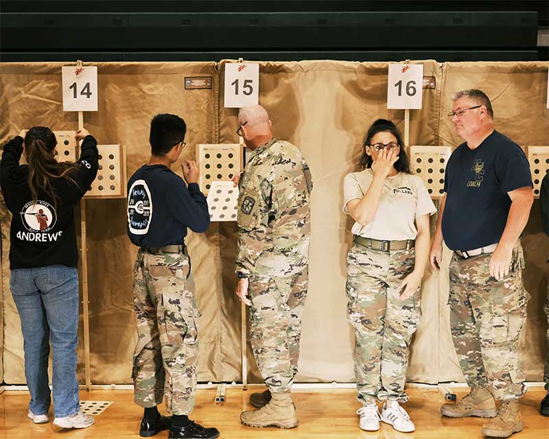 A line of students and instructors, some of them in camouflage uniforms, stand near a series of marksmanship target cards.